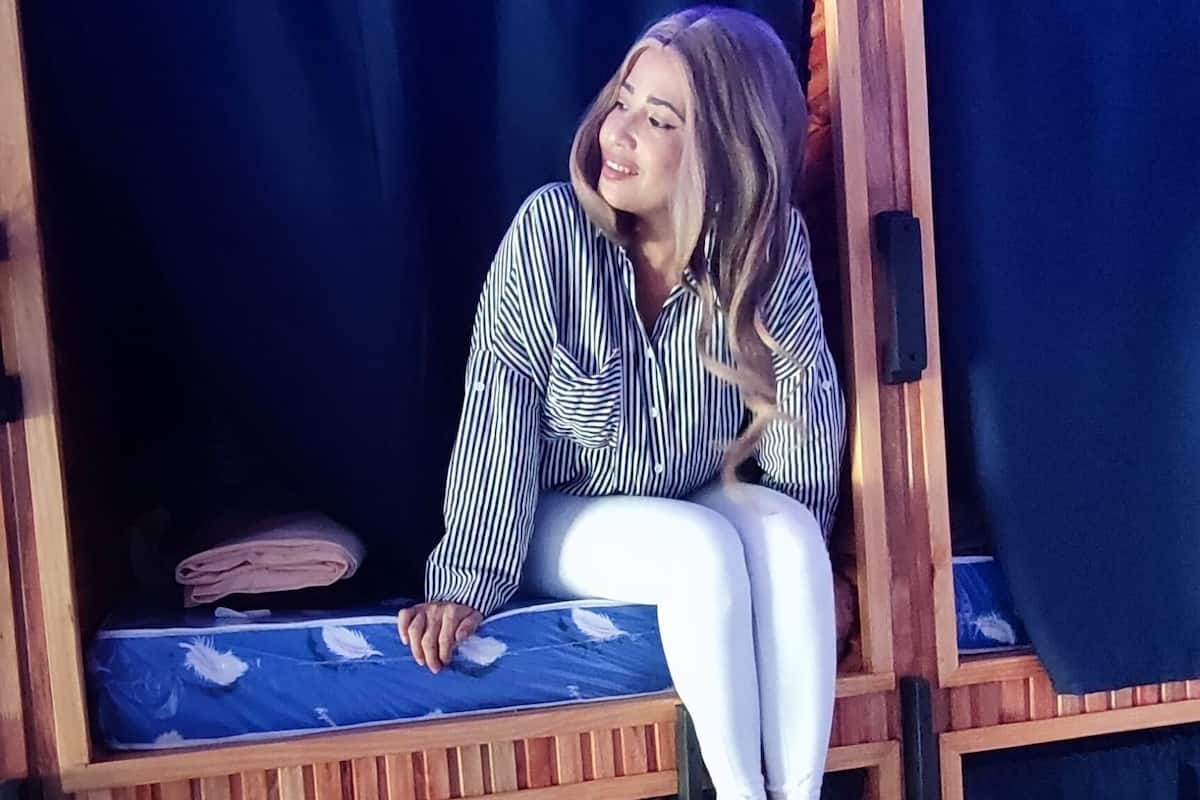 A person sitting on a bunk bed with a ladder, in a room with a dark curtain and a blue mattress.