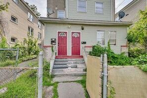 A two-story residential building with a red front door, a satellite dish, and a small front yard with a fence and some greenery.