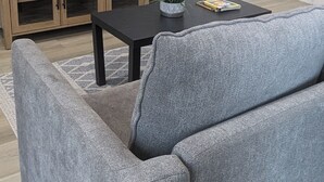 A living room with a grey sofa, a black coffee table, a wooden entertainment unit, a flat-screen TV, a white door, and a small plant on the table.
