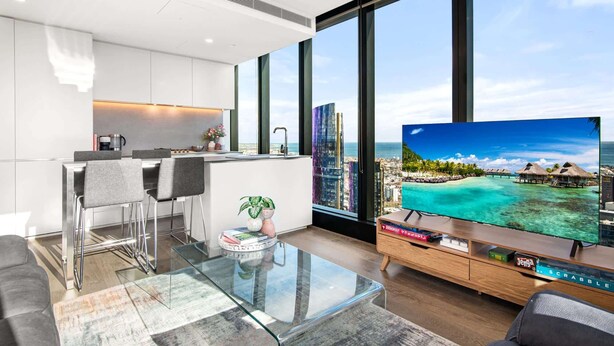 A modern living room with a large TV, a glass coffee table, and a view of a tropical beach and city skyline. - Stylish City Apartment with Expanding City Views (Southbank)