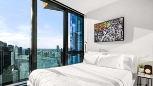 A modern bedroom with a large bed, a city view through floor-to-ceiling windows, and a colorful abstract painting on the wall. - Stylish City Apartment with Expanding City Views (Southbank)