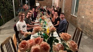 A group of people seated around a long table with a floral centerpiece, in an outdoor setting with brick walls and a window.