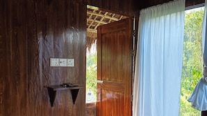Quarto de madeira com porta que dá para área externa, prateleira pequena com tomadas e vista para vegetação.