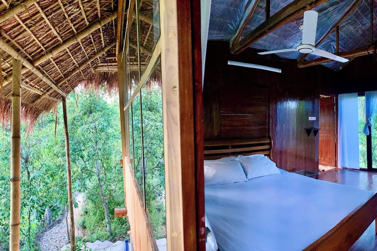 Una habitación con cama, ventilador de techo y amplios ventanales con vistas a un bosque.