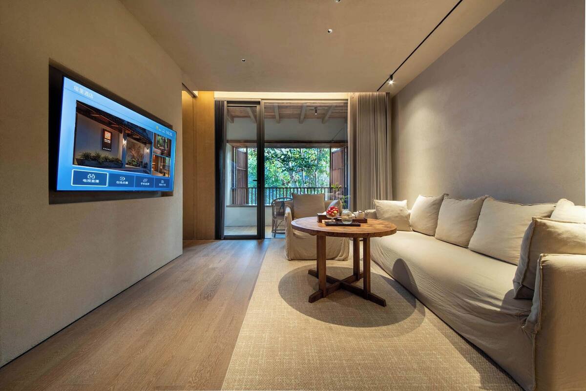 A modern hotel room with a large TV on the wall, a sofa, a round wooden table, and a view of the outdoors through a sliding door.