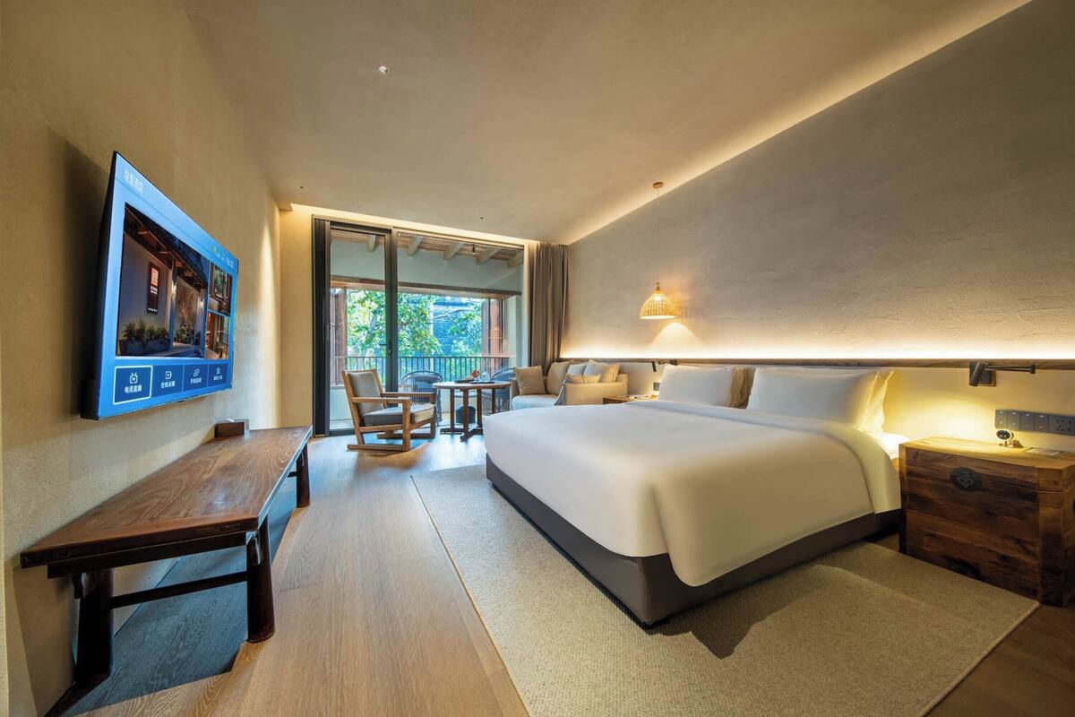 A modern hotel room with a large bed, a wooden bench, a desk, and a television mounted on the wall.