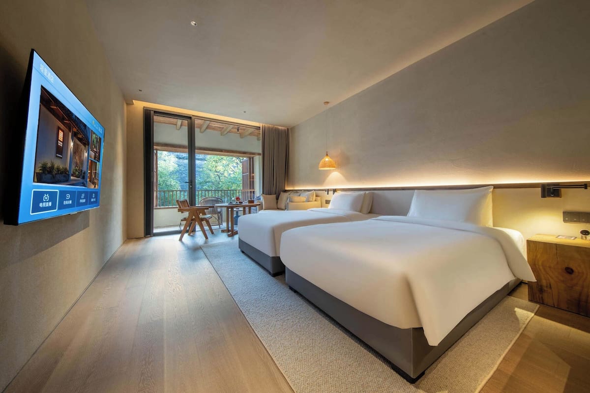 A modern hotel room with two beds, a TV mounted on the wall, a small table with chairs, and a large window offering a view of the outdoors.