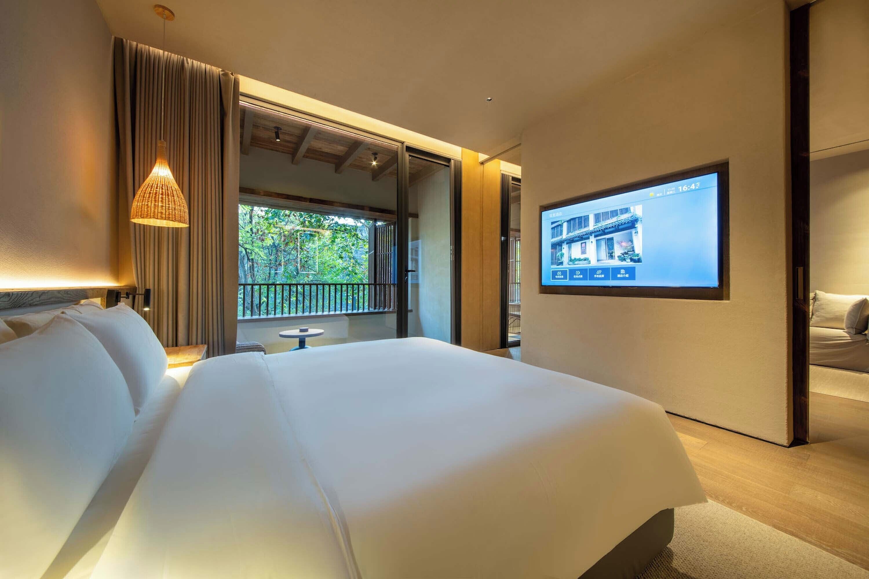 Habitación de hotel moderna con una cama grande, un televisor montado en la pared y un balcón con vistas a zonas verdes.