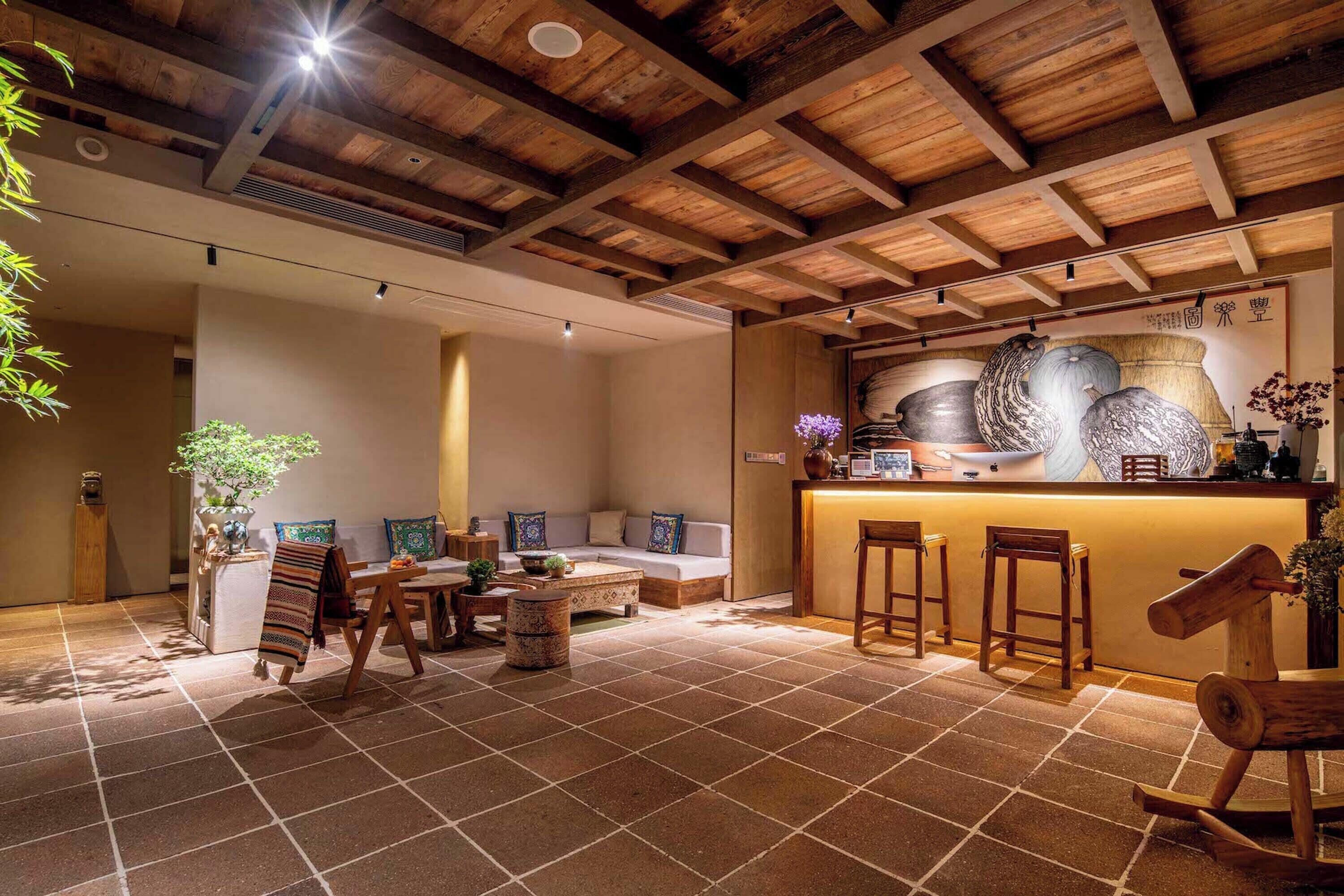 A cozy interior with a wooden ceiling, a bar area with stools, a seating area with a sofa and cushions, and a tiled floor.