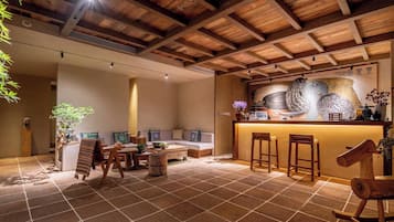A cozy interior with a wooden ceiling, a bar area with stools, a seating area with a sofa and cushions, and a tiled floor.