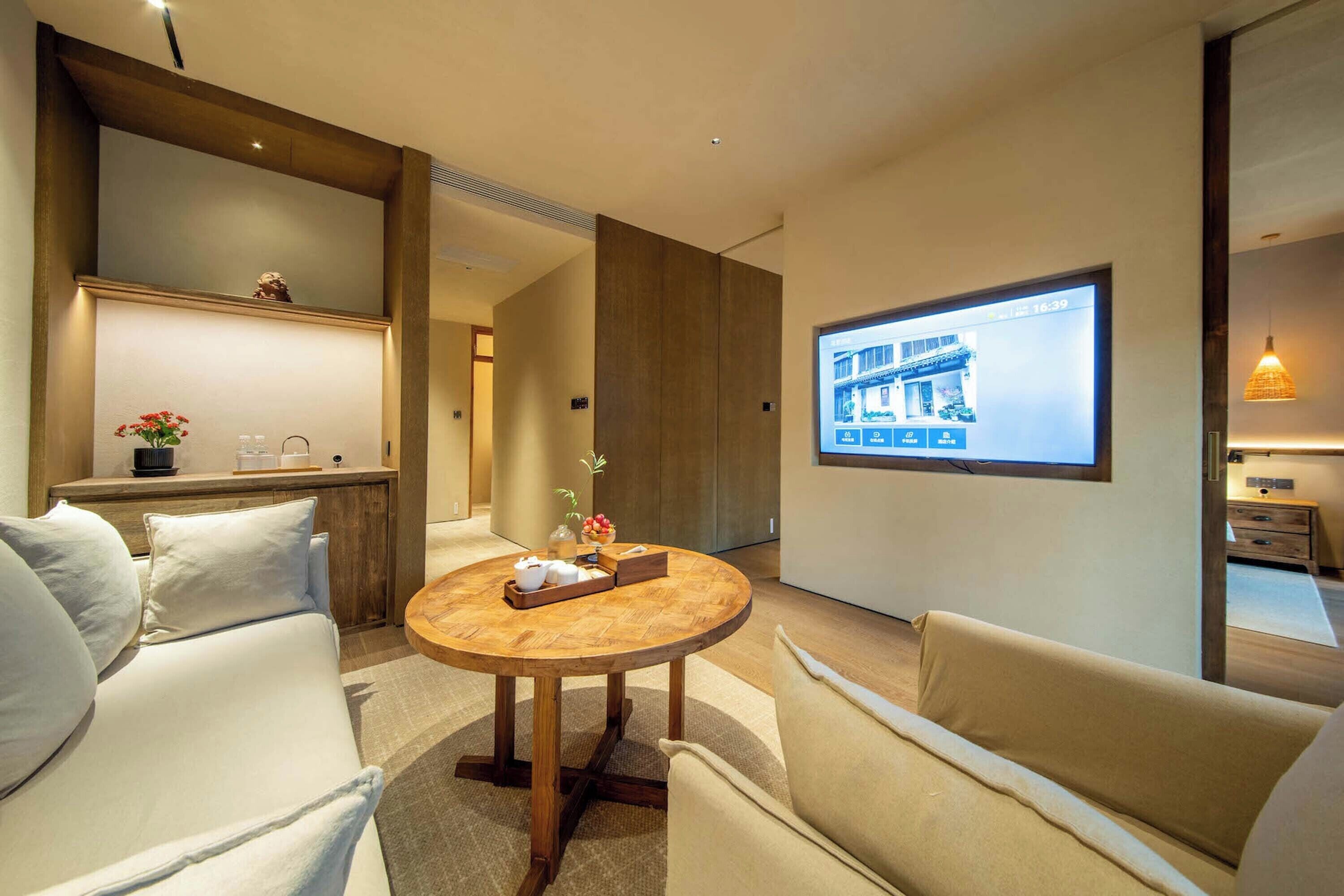Une chambre d’hôtel moderne avec une table ronde en bois, deux canapés, un écran de télévision affichant une image d’un bâtiment et un petit coin cuisine.