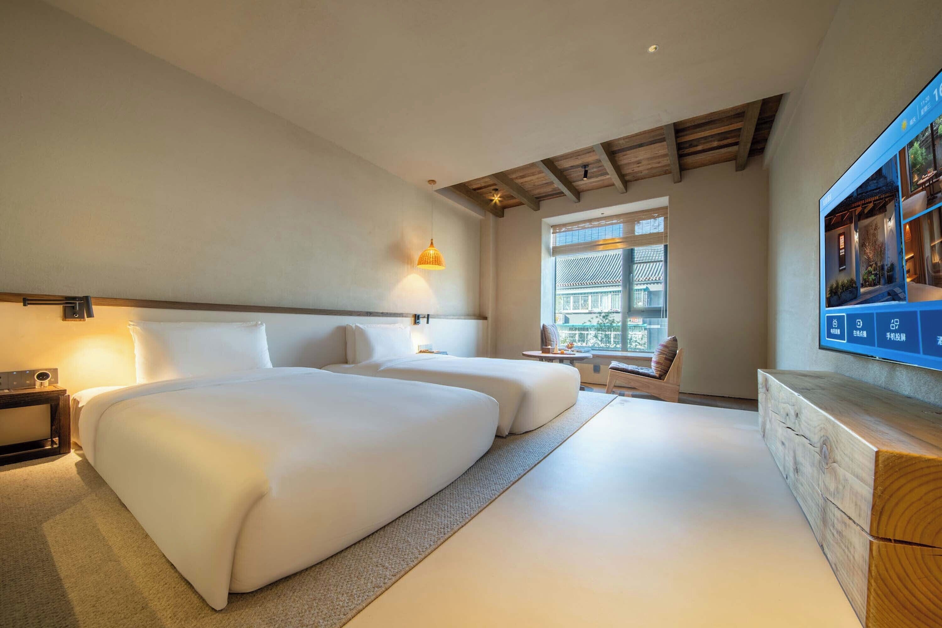 A hotel room with two beds, a wooden ceiling, a TV, and a window with blinds.