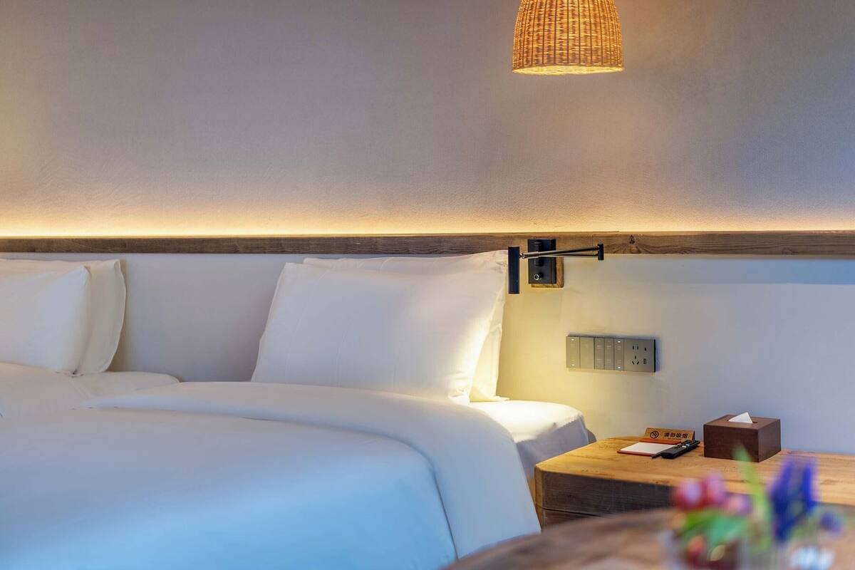 A neatly made bed with white linens, a bedside table with a lamp, and a wall-mounted power outlet.