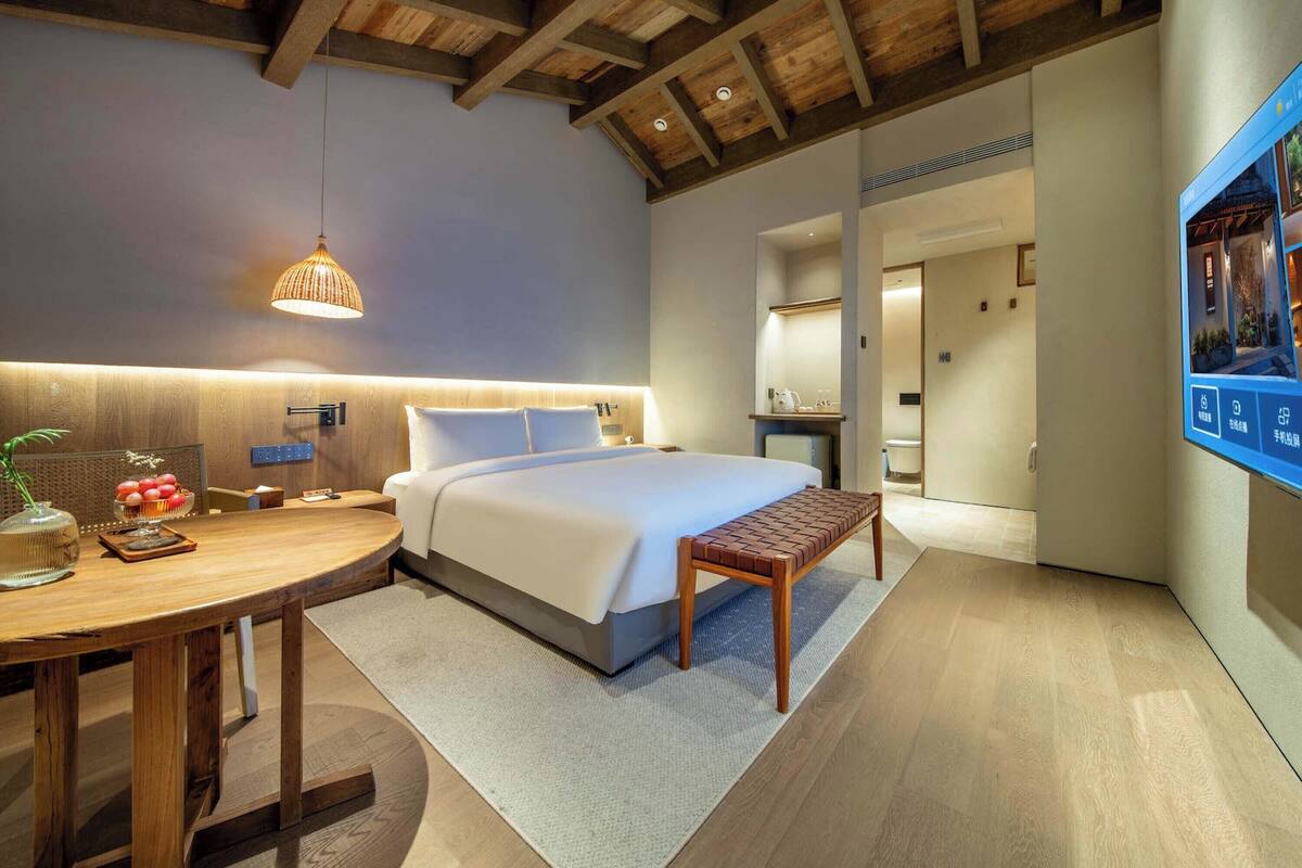 A modern hotel room with a large bed, a wooden table with a vase and fruit, a bench, and a wall-mounted TV.