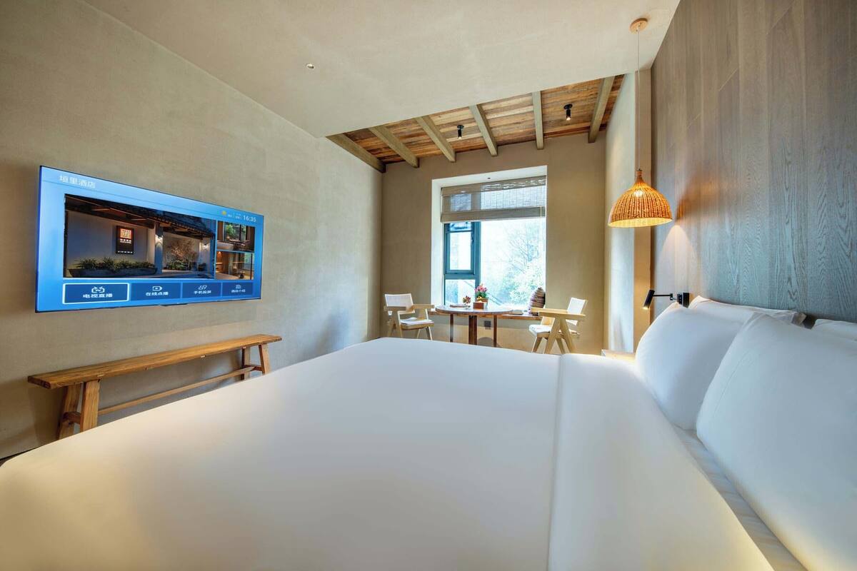 A modern hotel room with a large bed, a TV mounted on the wall, a wooden bench, a dining table with chairs, and a window with blinds.