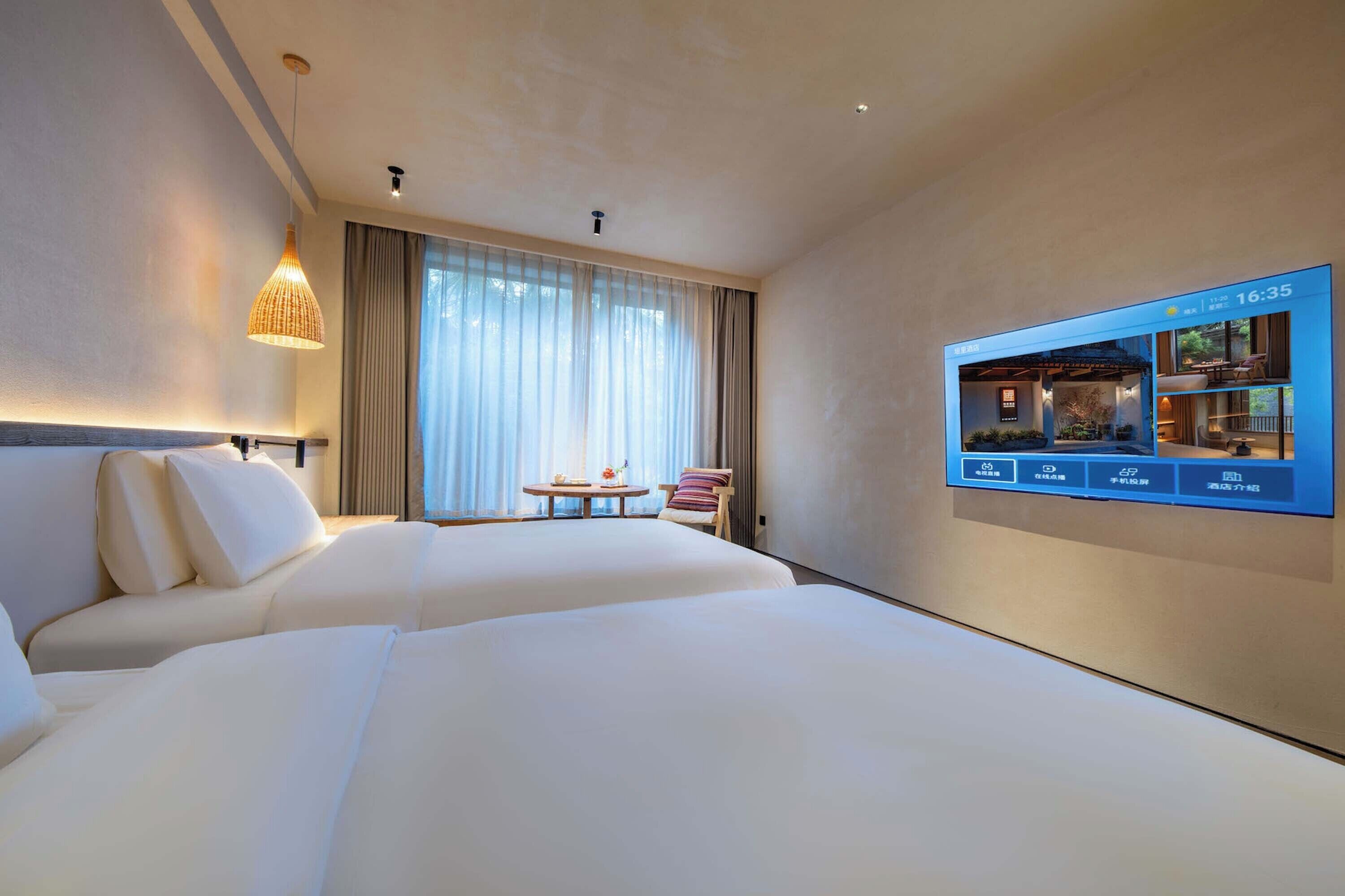 Une chambre d’hôtel avec deux lits, un téléviseur fixé au mur, une fenêtre avec des rideaux et une petite table avec une chaise.