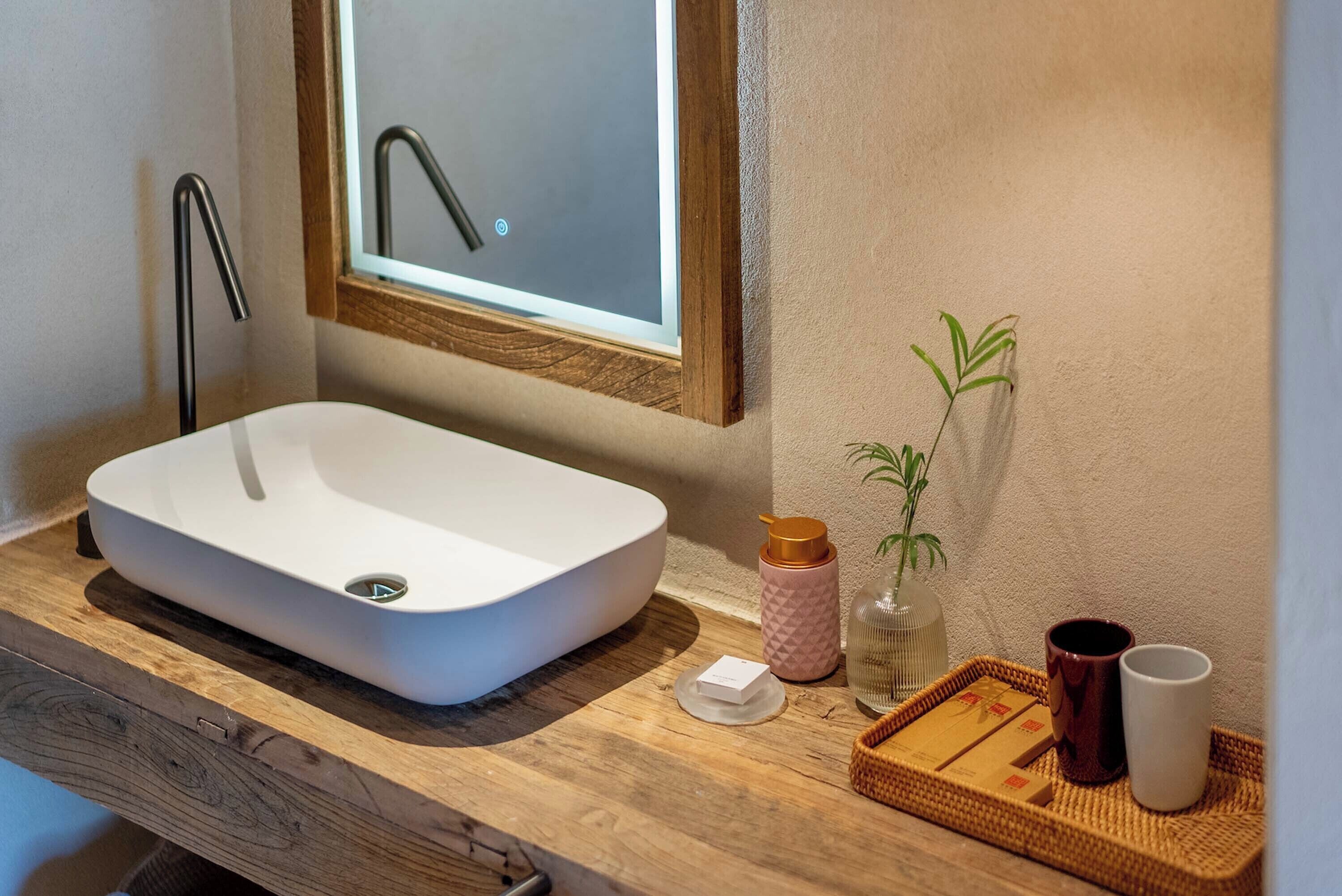 Une salle de bain avec un lavabo blanc, un meuble-lavabo en bois, un miroir, une plante en pot, une bouteille et un plateau sur lequel se trouvent une tasse et une boîte.