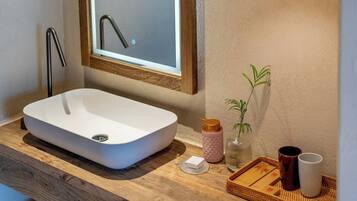 Une salle de bain avec un lavabo blanc, un meuble-lavabo en bois, un miroir, une plante en pot, une bouteille et un plateau sur lequel se trouvent une tasse et une boîte.