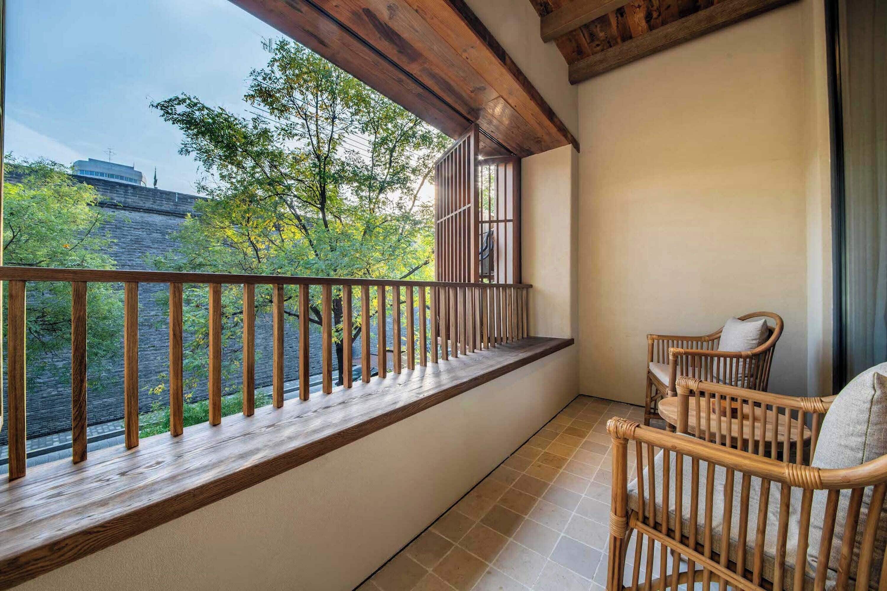 Een balkon met een houten hekwerk, uitzicht op bomen en een gebouw, en twee rieten stoelen.