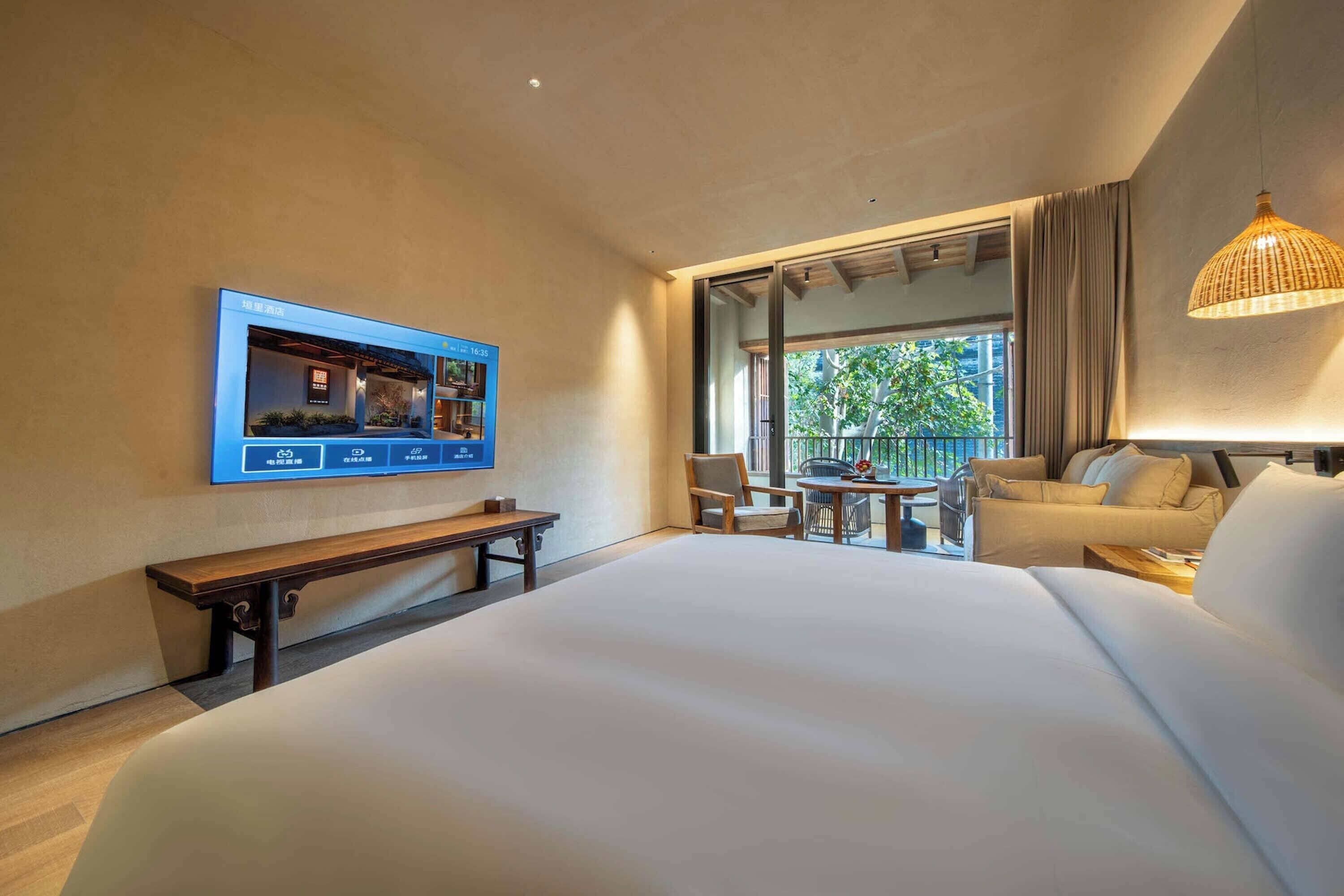 Habitación de hotel con una cama grande, un televisor montado en la pared, una mesita con sillas y un balcón con vistas a zonas verdes.