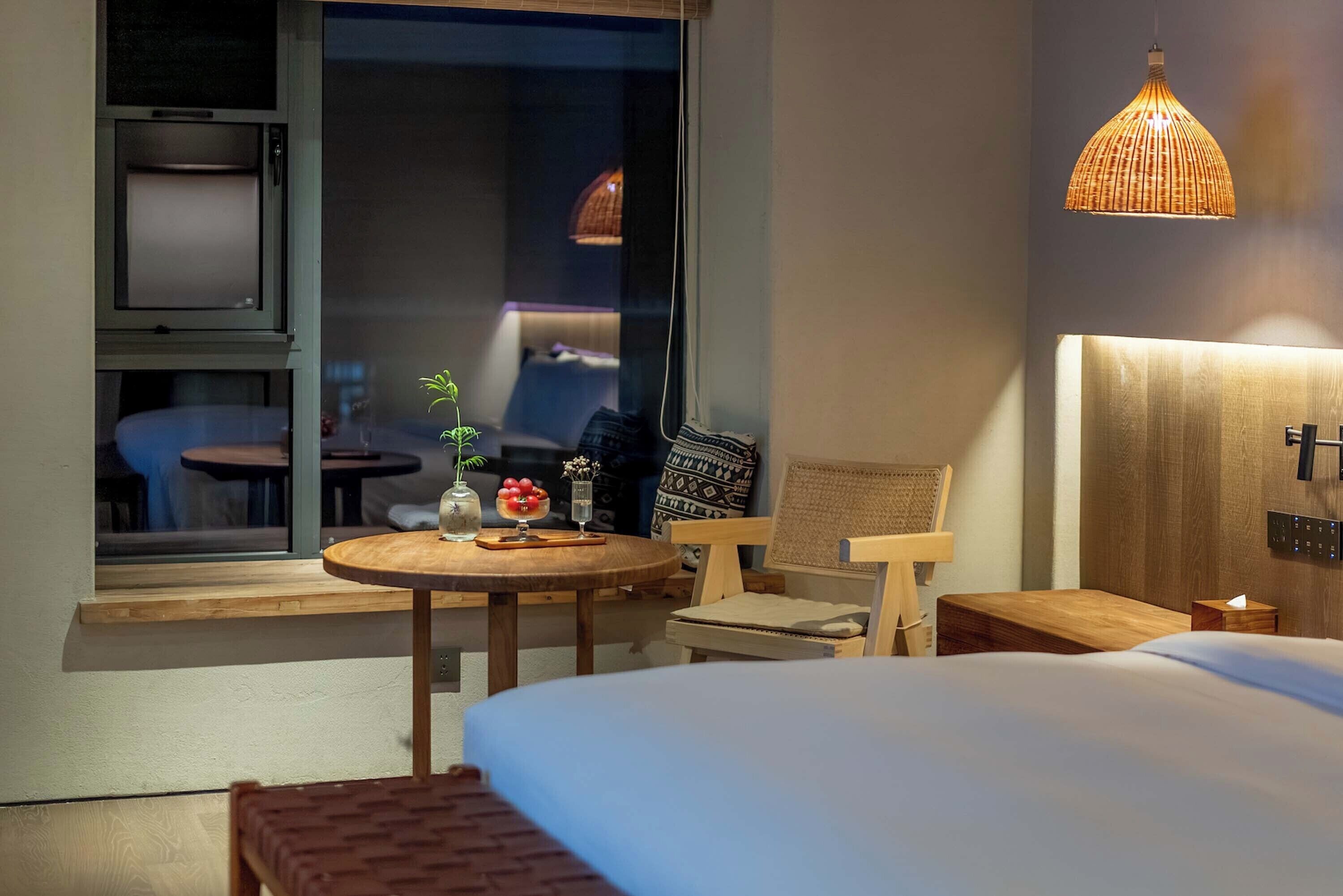 Een hotelkamer met een bed, een klein rond tafeltje met een vaas en fruit, een stoel en een raam met uitzicht.