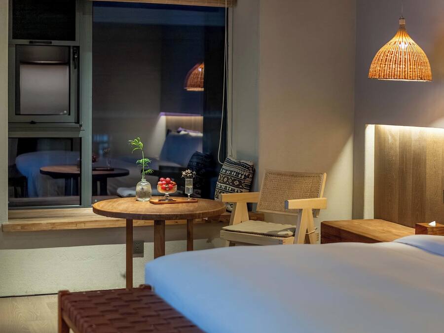 Een hotelkamer met een bed, een klein rond tafeltje met een vaas en fruit, een stoel en een raam met uitzicht.