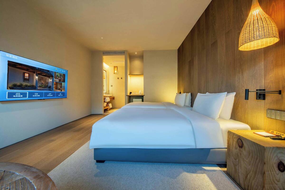 A modern hotel room with a large bed, a wooden accent wall, a mounted television, and a bedside table with a lamp.