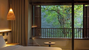 Une chambre d’hôtel avec un grand lit, une petite table ronde, une lampe et une vue sur la verdure par une fenêtre donnant sur un balcon.