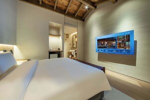 Habitación de hotel con una cama grande, una mesita de noche, un espejo y un televisor montado en la pared mostrando la distribución de la habitación.