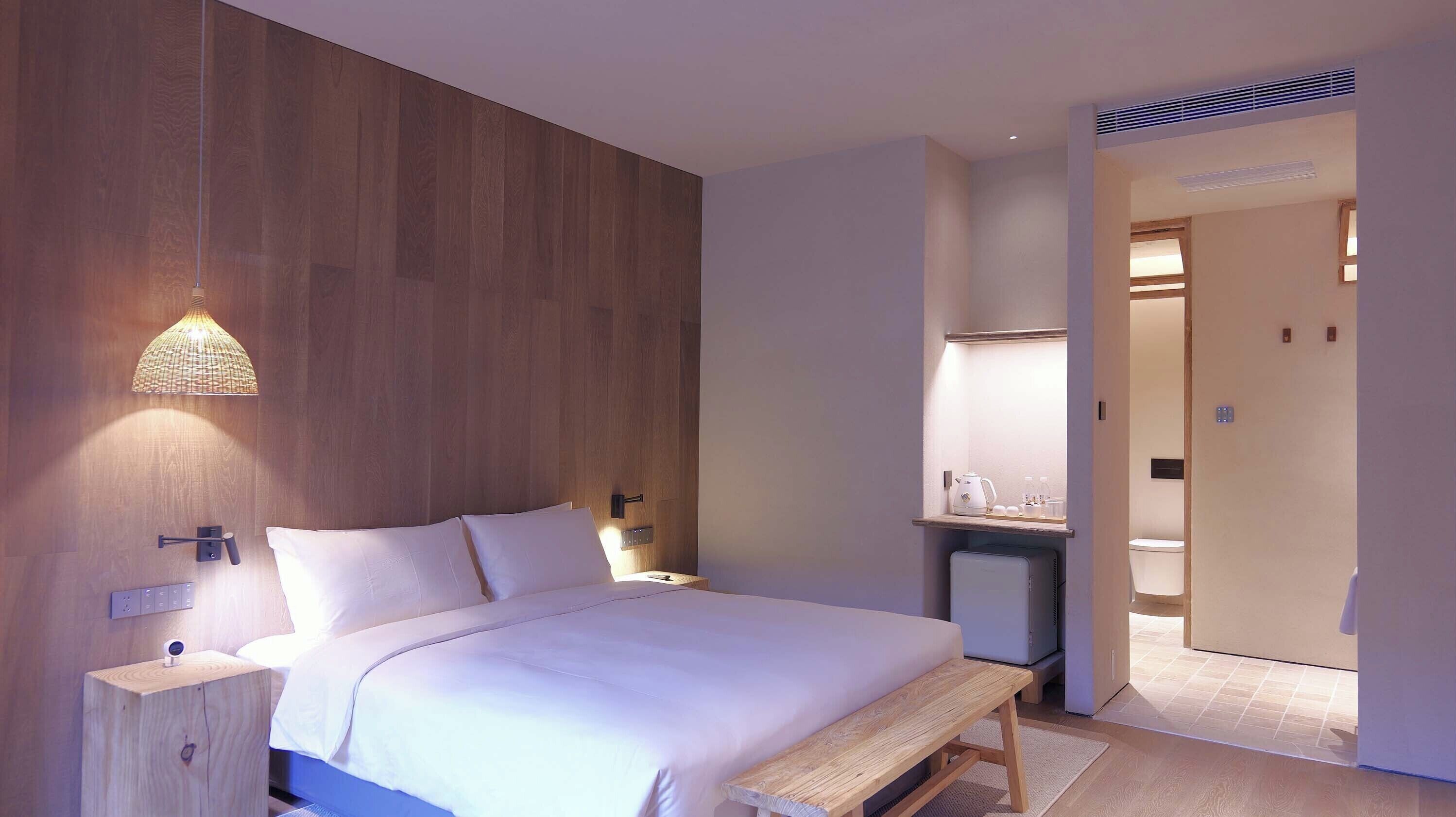Una habitación de hotel moderna con un revestimiento de pared de madera, una cama con ropa blanca, una mesita de noche con lámpara y un pequeño banco.