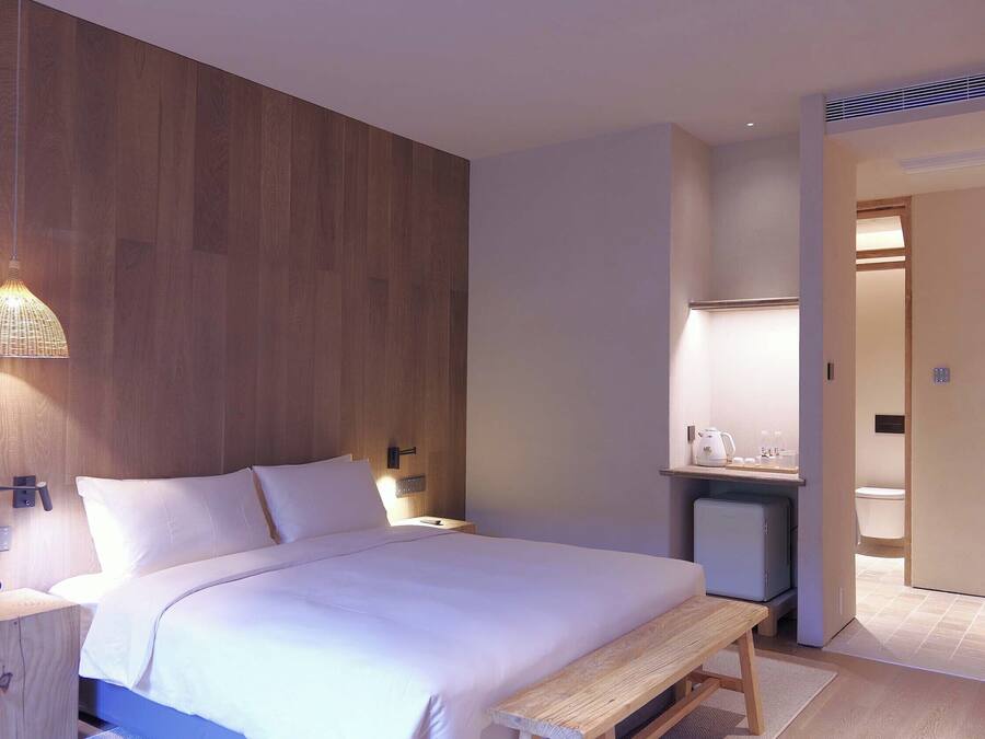 Una habitación de hotel moderna con un revestimiento de pared de madera, una cama con ropa blanca, una mesita de noche con lámpara y un pequeño banco.