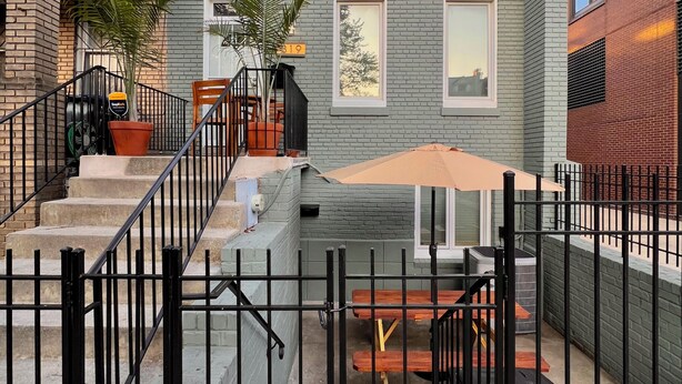 Edificio de dos pisos con fachada de ladrillo, un pequeño patio con mesa y sillas, y una cerca de metal negro.