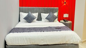 Una cama con cabecera gris, ropa de cama blanca y almohadas estampadas. Se observa una cortina roja detrás de la cama y una mesita de noche con un teléfono y una lámpara.