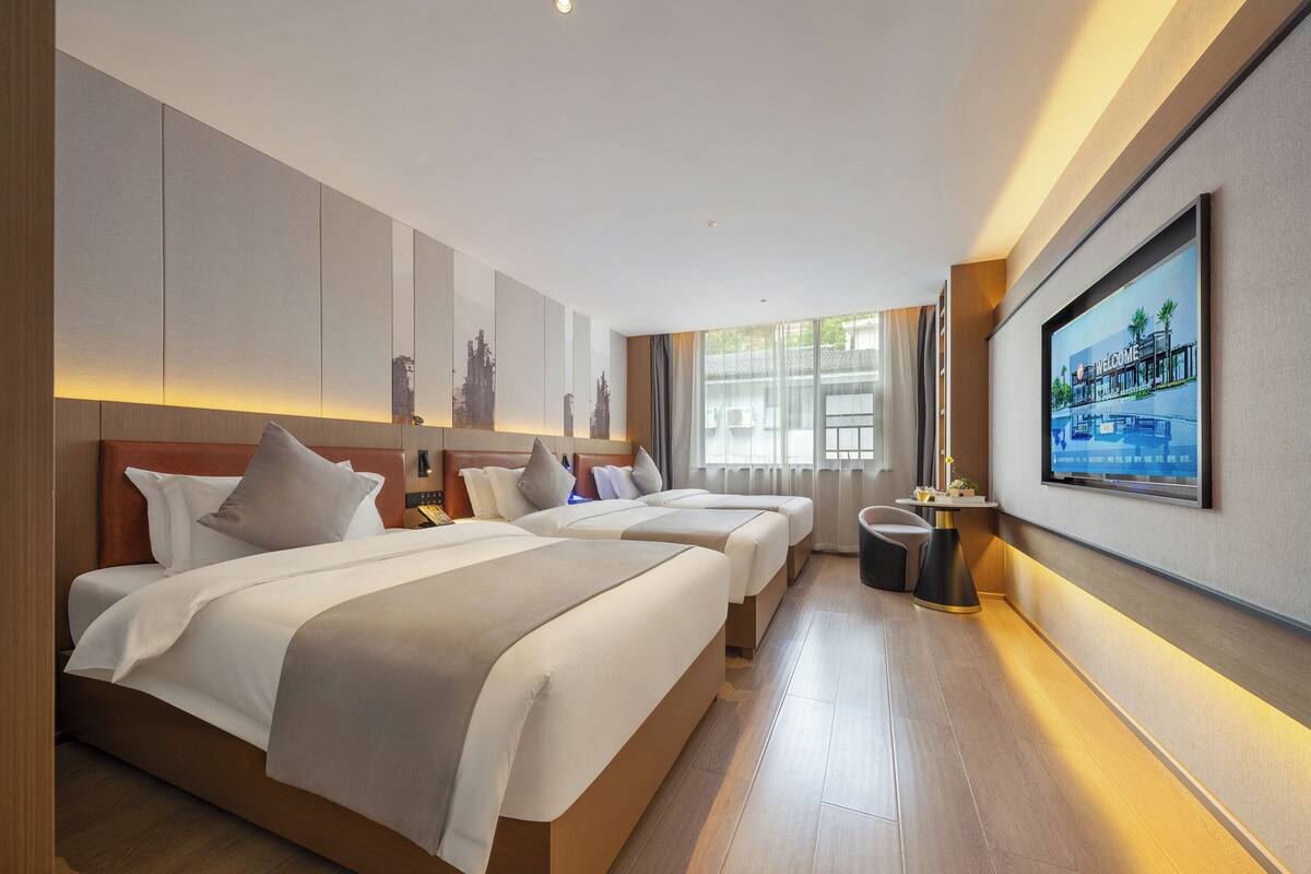 Habitación de hotel con dos camas, un ventanal grande, un televisor montado en la pared y un cartel de bienvenida enmarcado.