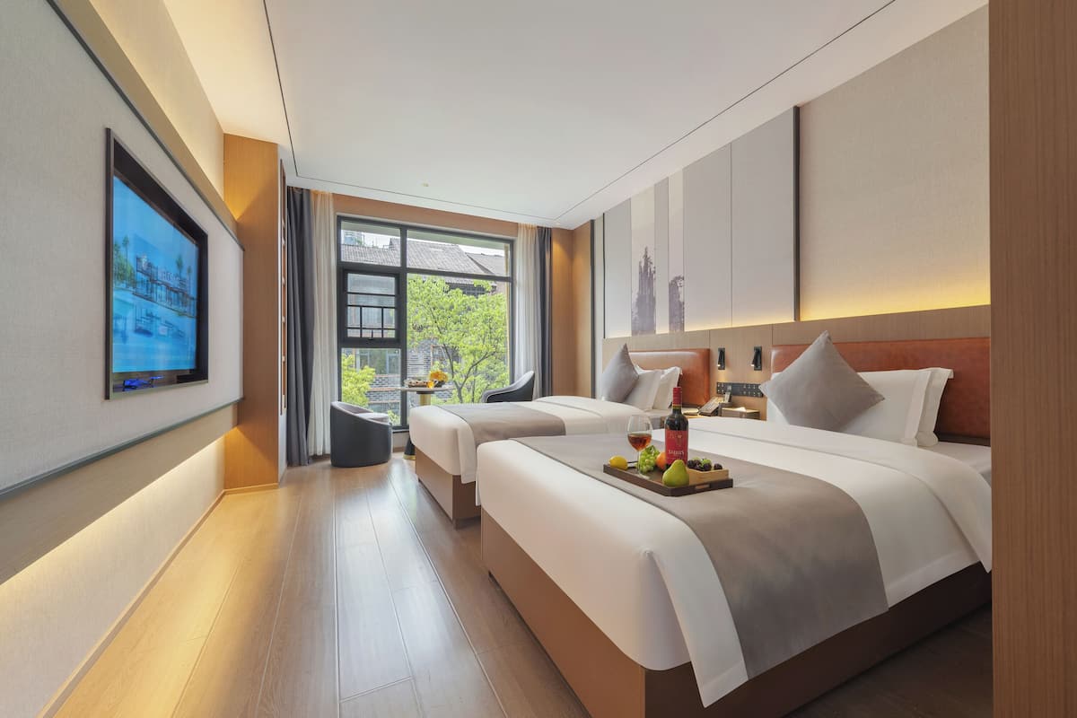 Una habitación de hotel moderna con una cama grande, una cama más pequeña, un televisor y una ventana con vistas a zonas verdes.