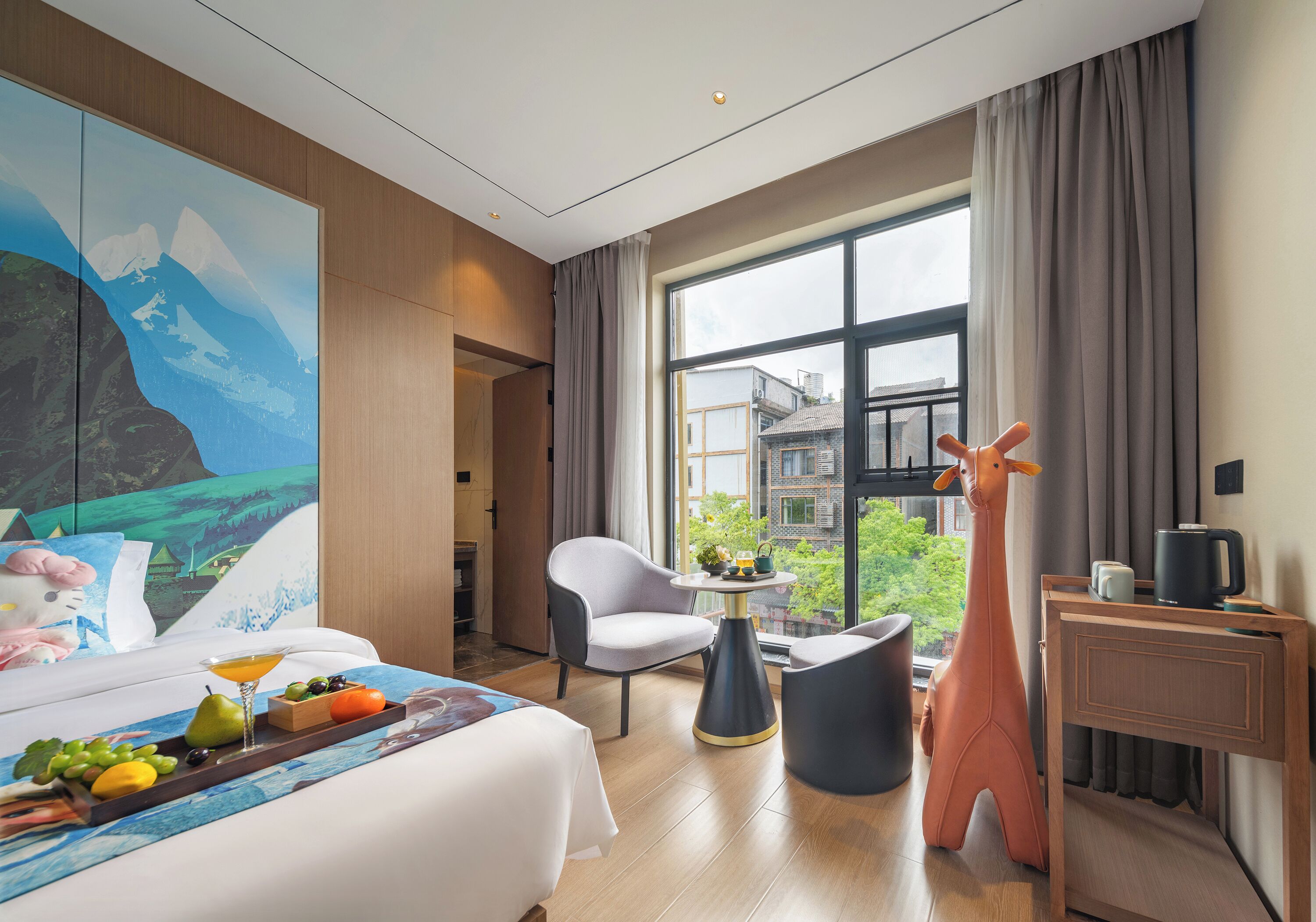 A hotel room with a bed, a desk, a chair, a giraffe statue, and a large window with a view of buildings and greenery.