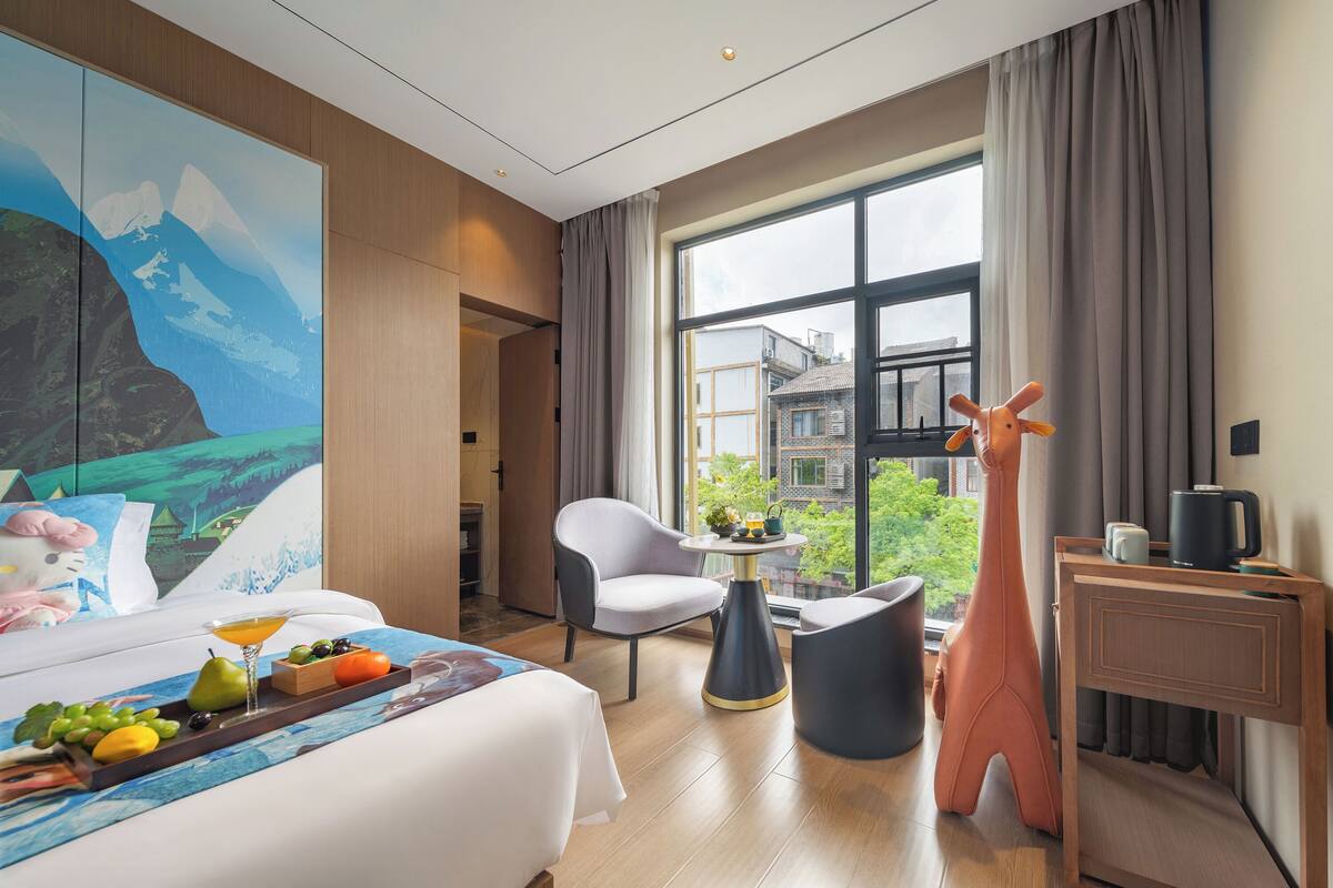 Habitación de hotel con cama, escritorio, silla, estatua de jirafa y un ventanal con vistas a edificios y follaje.