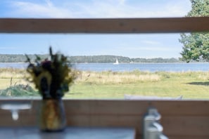 Blick durch ein Fenster mit Holzrahmen, eine Vase mit Blumen und ein Segelboot in der Ferne auf dem Wasser.
