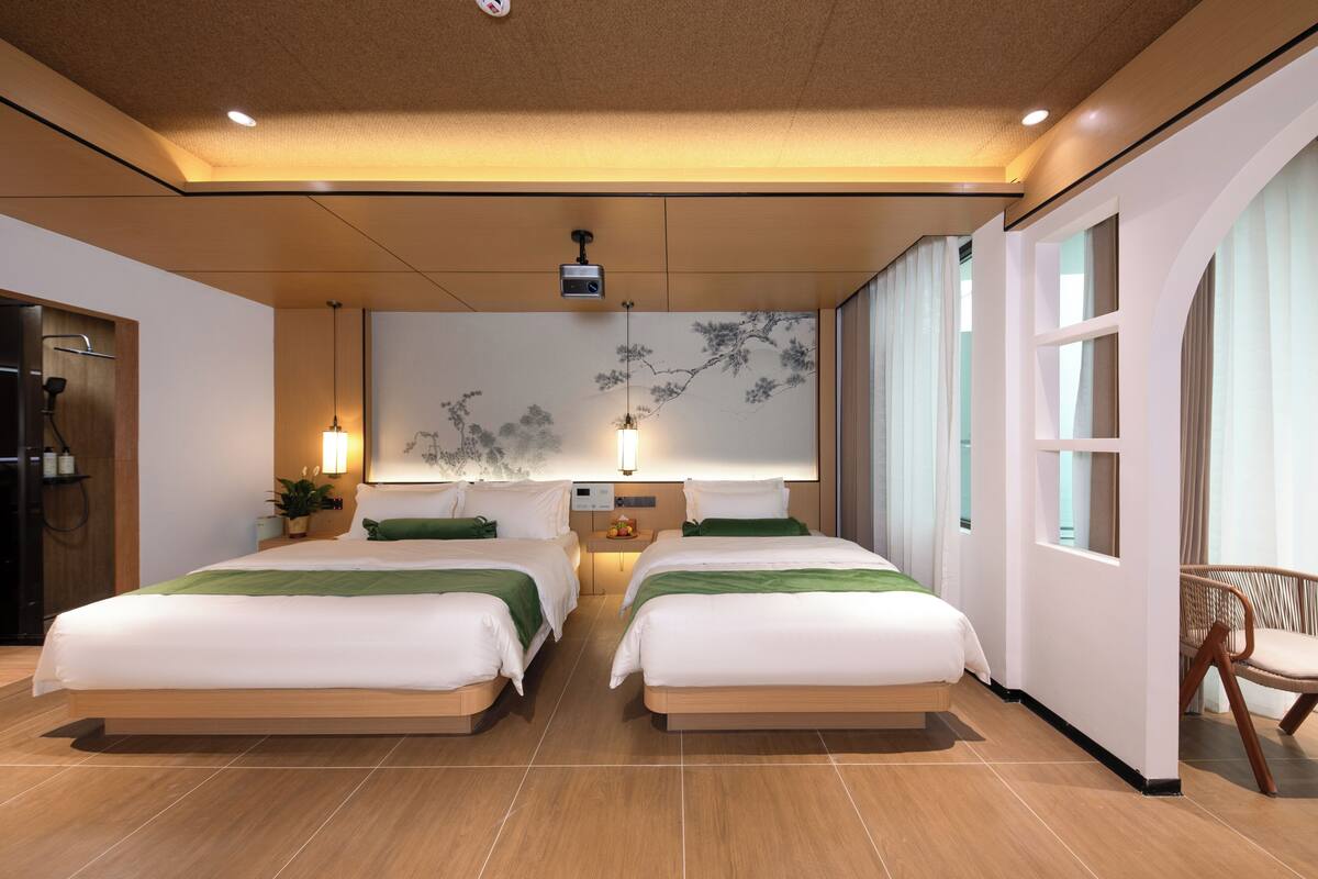 Habitación de hotel moderna con dos camas, un ventanal grande, un proyector montado en la pared y una pequeña mesa con una planta.