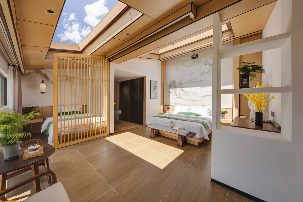 Una habitación de hotel moderna con suelo de madera, cama con cubrecama verde y blanco, zona de comedor con mesa y sillas, y vista al cielo a través de un tragaluz.