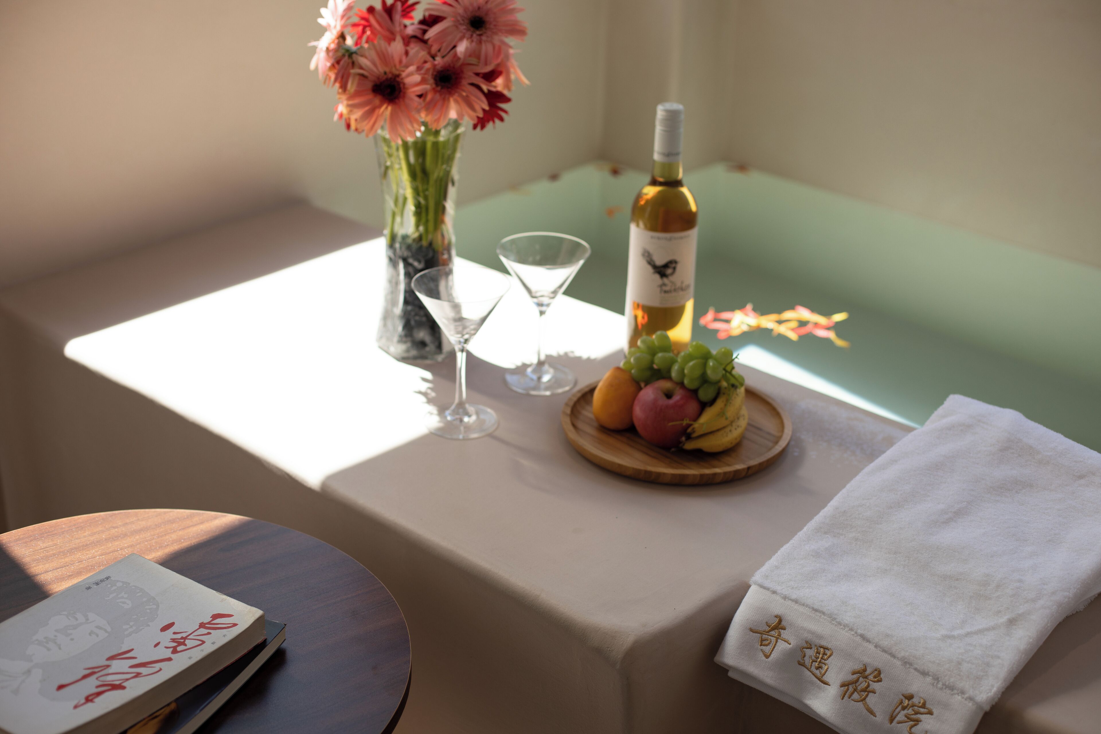 Una mesa con un libro, un jarrón con flores, una botella de vino, una copa de martini y un plato de fruta.