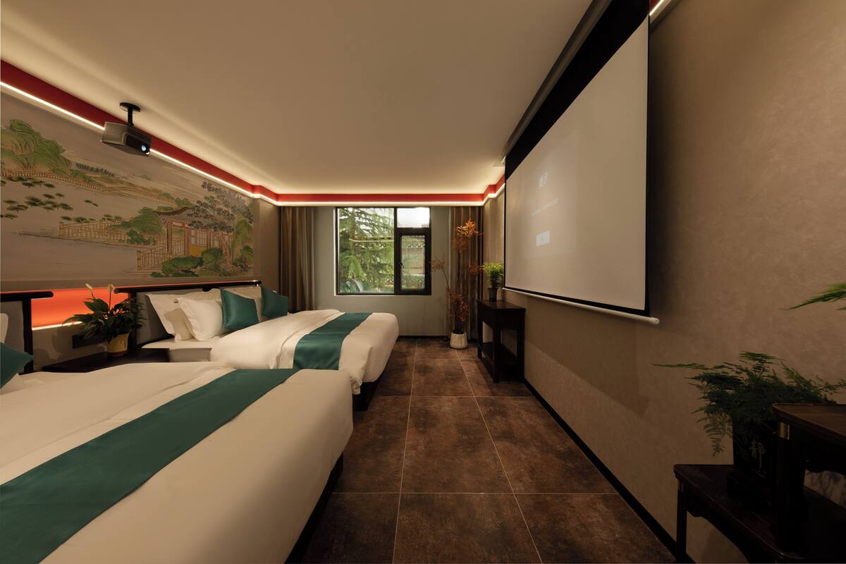 Una habitación de hotel con dos camas, un ventanal grande, una pantalla de proyector y un mural en la pared.