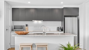 A modern kitchen with grey cabinets, a white countertop, a built-in microwave, a sink, and a refrigerator. There are two stools and a plant in the foreground. - Waterside Victor Harbor (Victor Harbor)