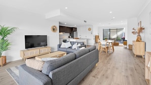 A modern living room with a grey sofa, a flat-screen TV, and a dining area with a table and chairs. - Waterside Victor Harbor (Victor Harbor)