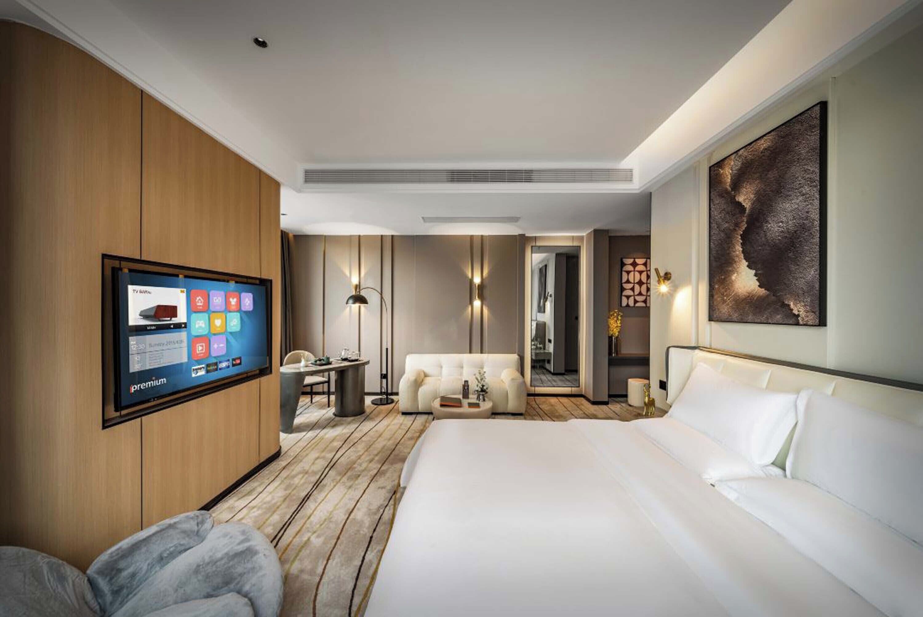 A modern hotel room with a large bed, a flat-screen TV mounted on the wall, a sofa, a small table, and a decorative wall piece.