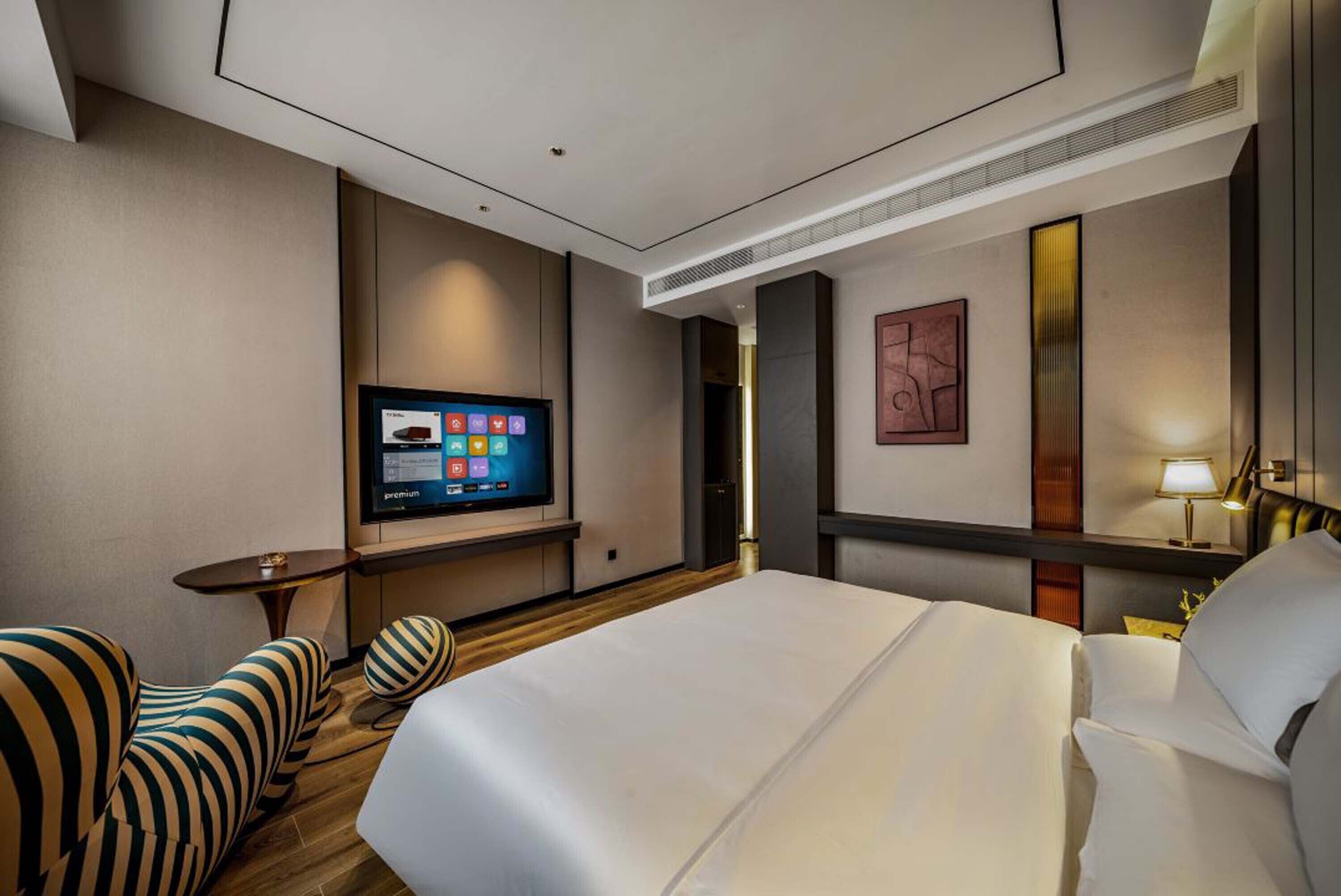 Een hotelkamer met een groot bed, een televisie aan de muur, een klein tafeltje en gestreepte fauteuils.