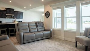A living room with a gray sofa, a dining area with a kitchen in the background, and large windows providing natural light.