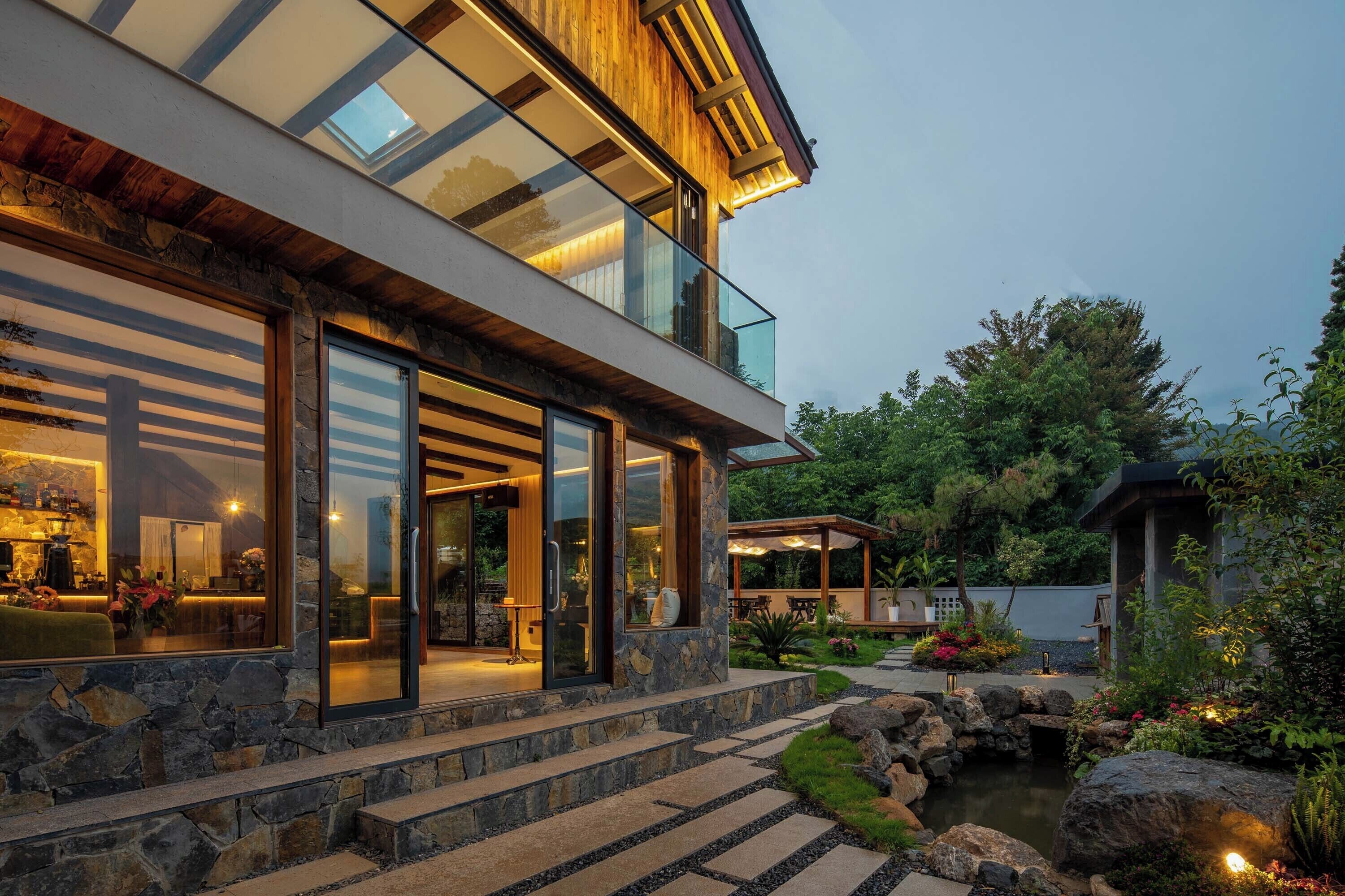 A modern house with a stone facade, large glass windows, and a well-lit outdoor area with a pond and garden.