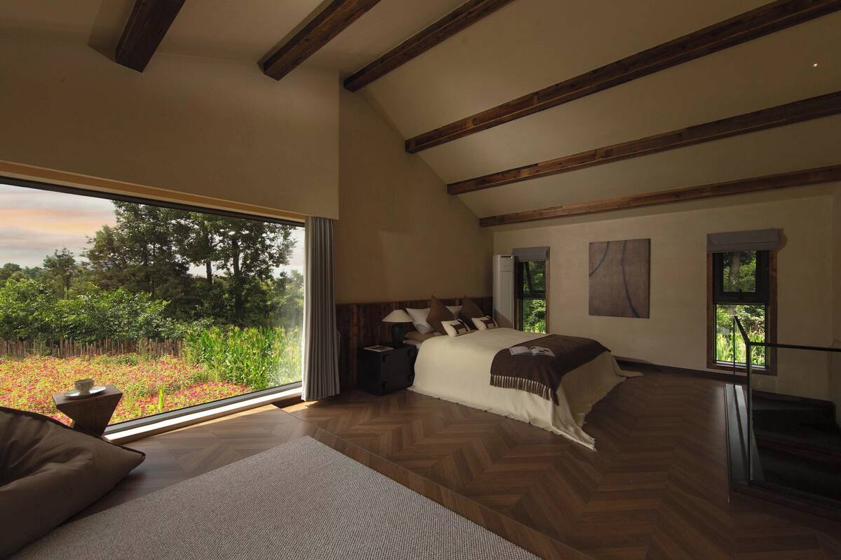 A bedroom with a large window, wooden beams, a bed, and a view of greenery.