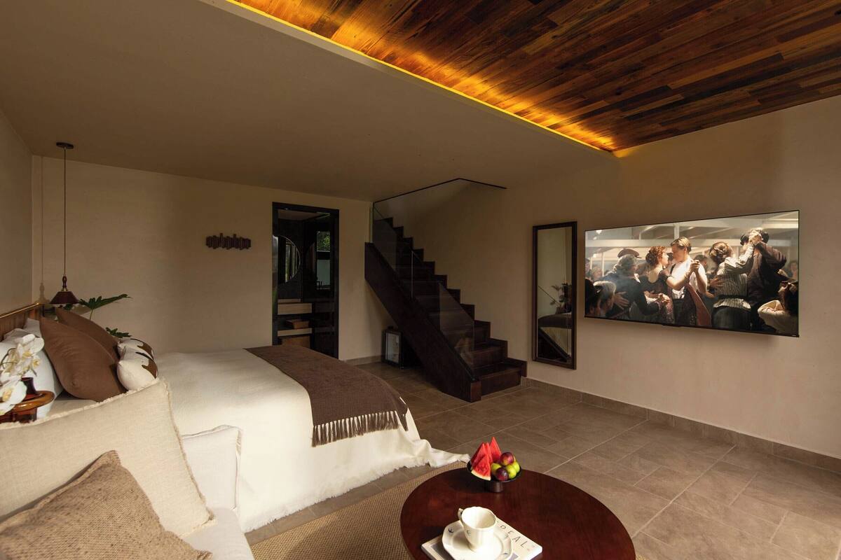 A modern hotel room with a bed, a coffee table with fruit, a painting, and a staircase.